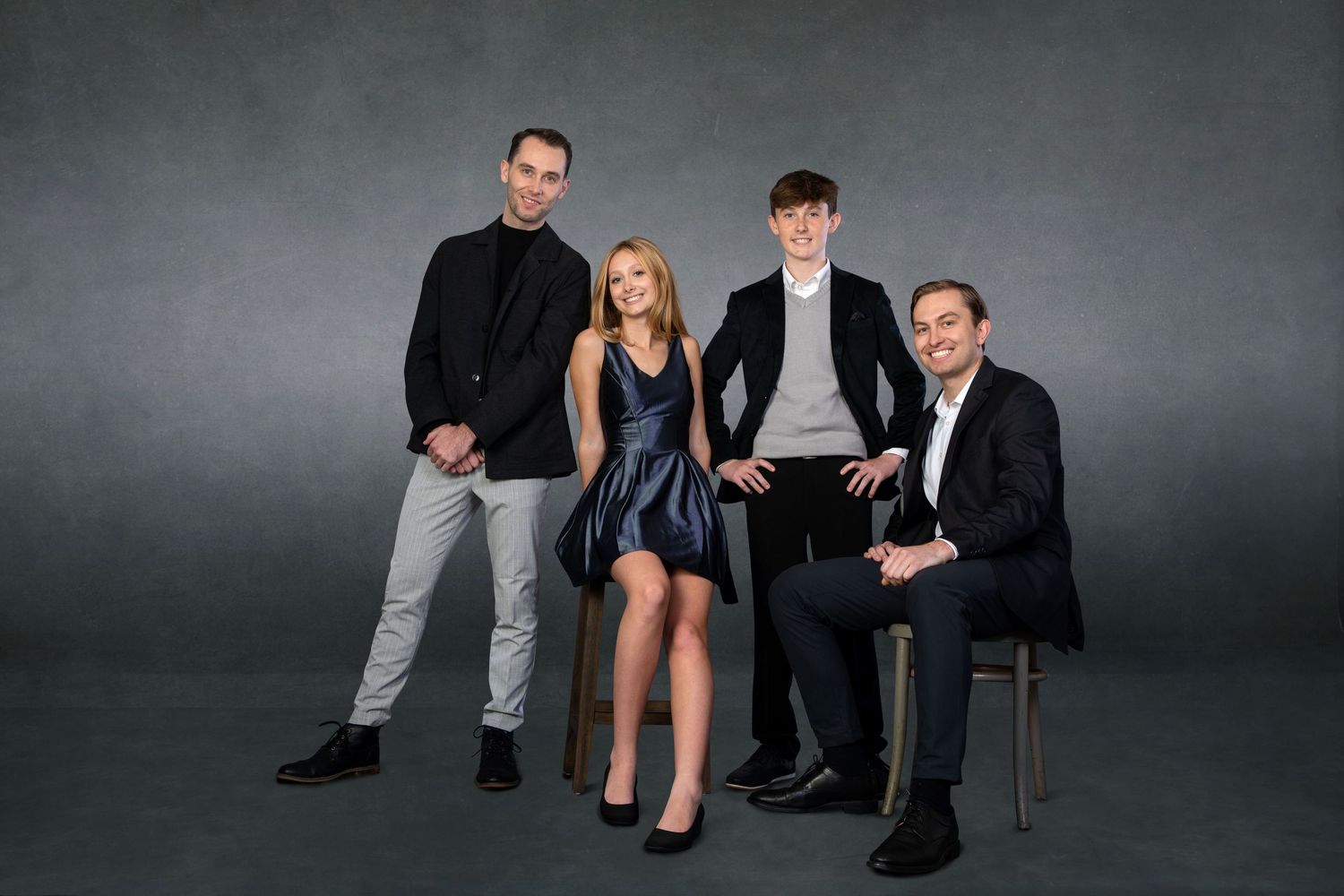 Four elegantly dressed people pose for a professional studio portrait against a gray background.