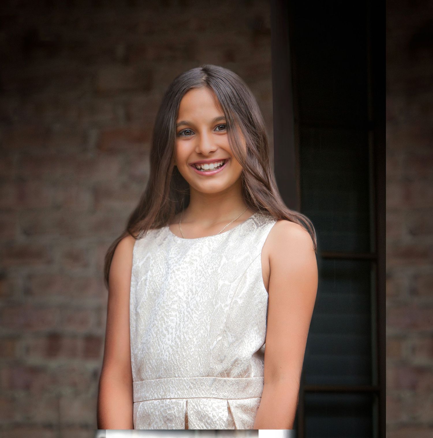 Smiling young girl with long brown hair wearing a white sleeveless dress, posing outdoors near a brick wall.