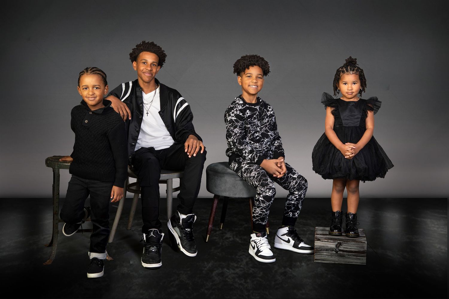 Four children dressed in stylish black outfits pose together against a dark gray studio background.