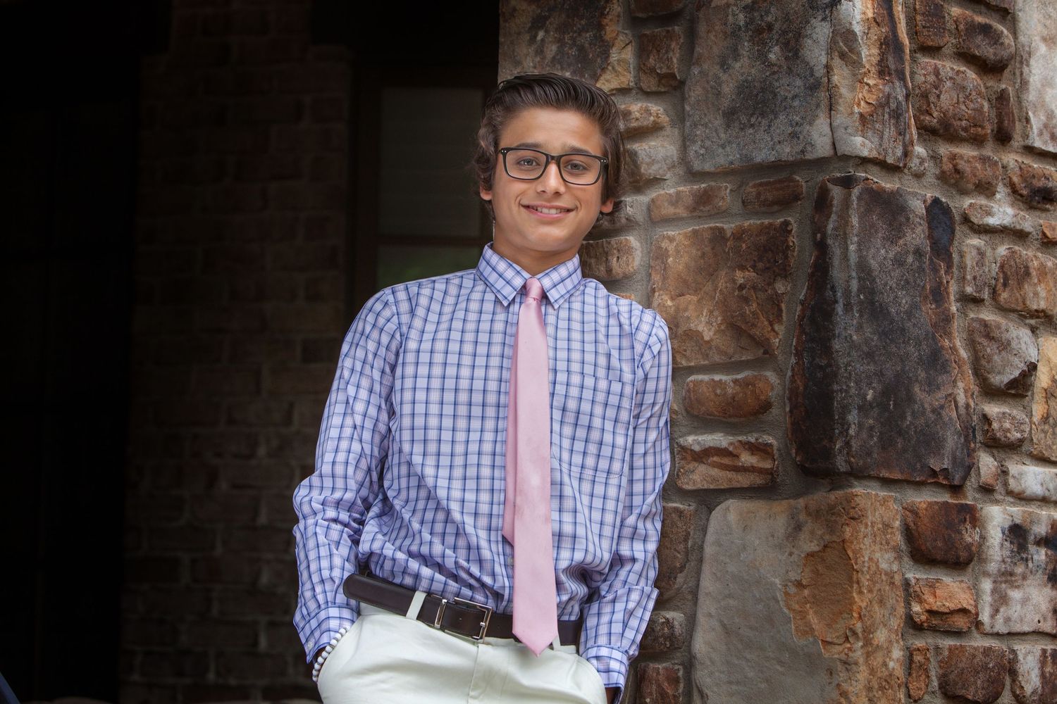 Young man in plaid shirt and pink tie smiling outdoors near stone wall.