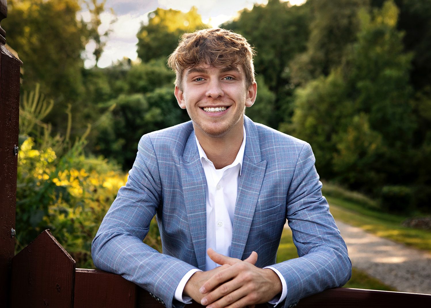 Smiling senior boy in blue plaid blazer poses outdoors with green trees and yellow flowers, by Jocelyn M. Photography in Rochester, MI