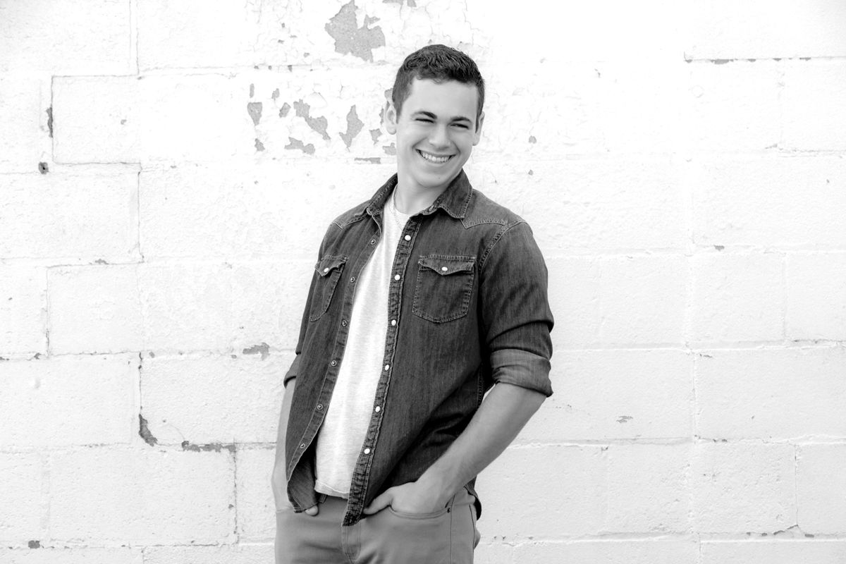Young man smiling confidently in denim shirt against white brick wall, black and white portrait photo.