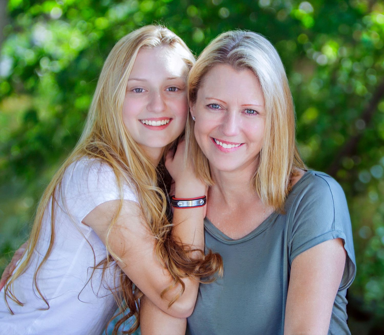 Two smiling blonde women pose together outdoors with lush green trees in the background.