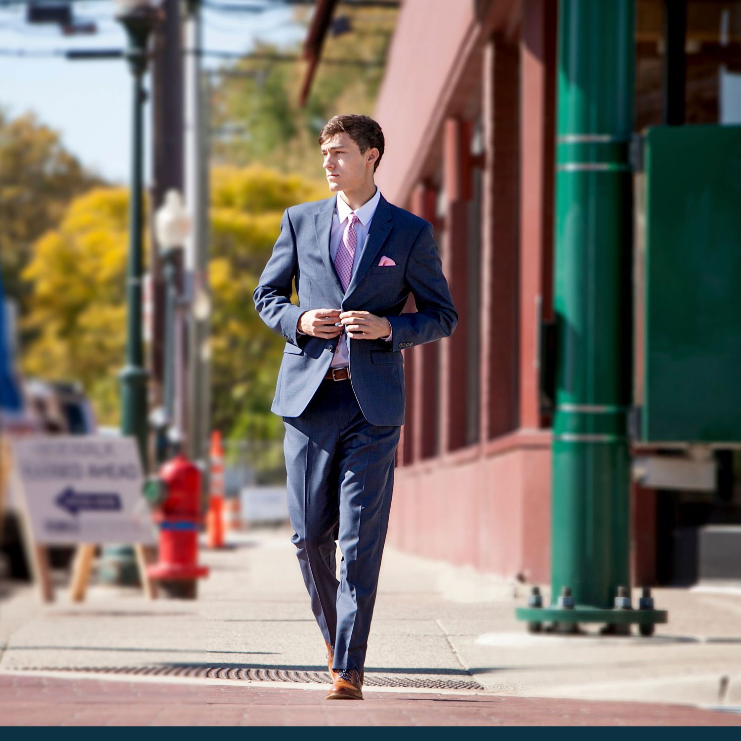 Young man in navy blue suit walks confidently down urban sidewalk for formal portrait.