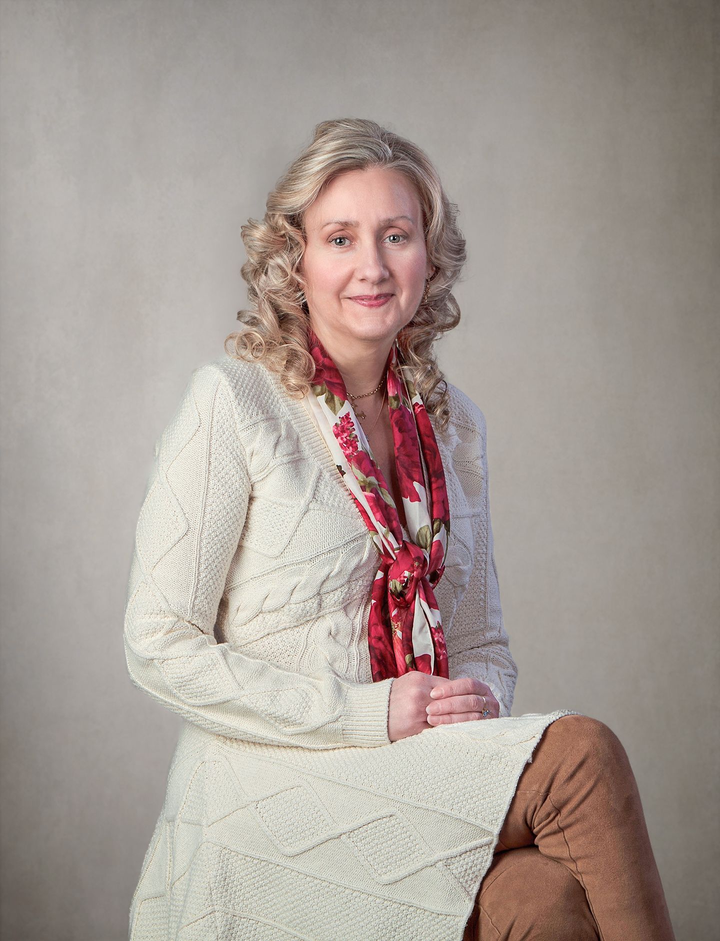 Elegant blonde woman in cream cable-knit sweater with floral scarf seated against neutral backdrop.