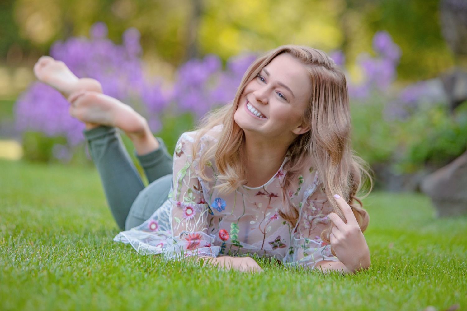 Young blonde woman smiling while lying on lush green grass surrounded by purple flowers in a garden.