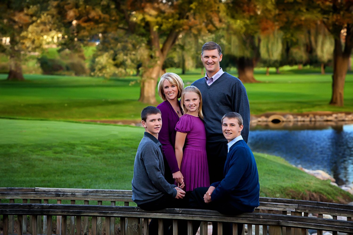 Family of five posing on a wooden bridge by a pond in a lush green golf course setting during autumn.