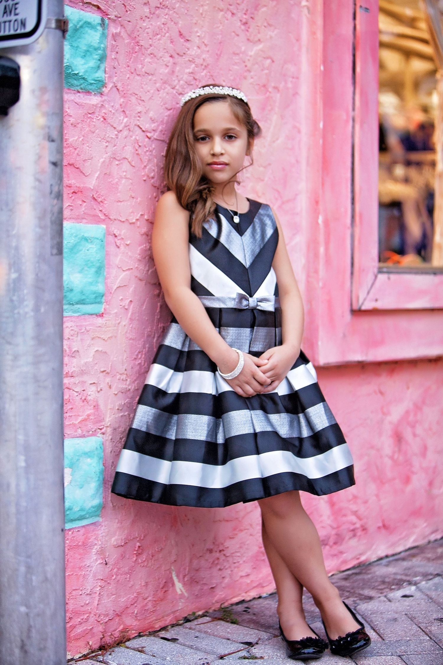 Young girl in black and white striped dress with bow, wearing headband, posing against pink wall in Rochester, MI by Jocelyn M. Photography