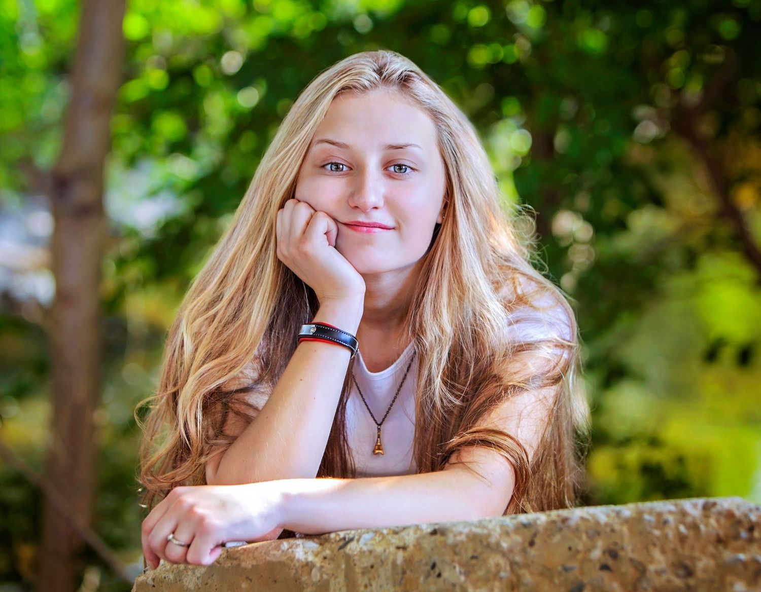 Young blonde teenage girl resting chin on hand, leaning on stone wall outdoors with green trees in background.