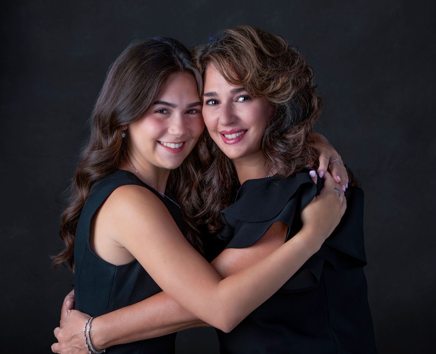 Two young women in black outfits share a warm embrace, smiling in a professional studio portrait.