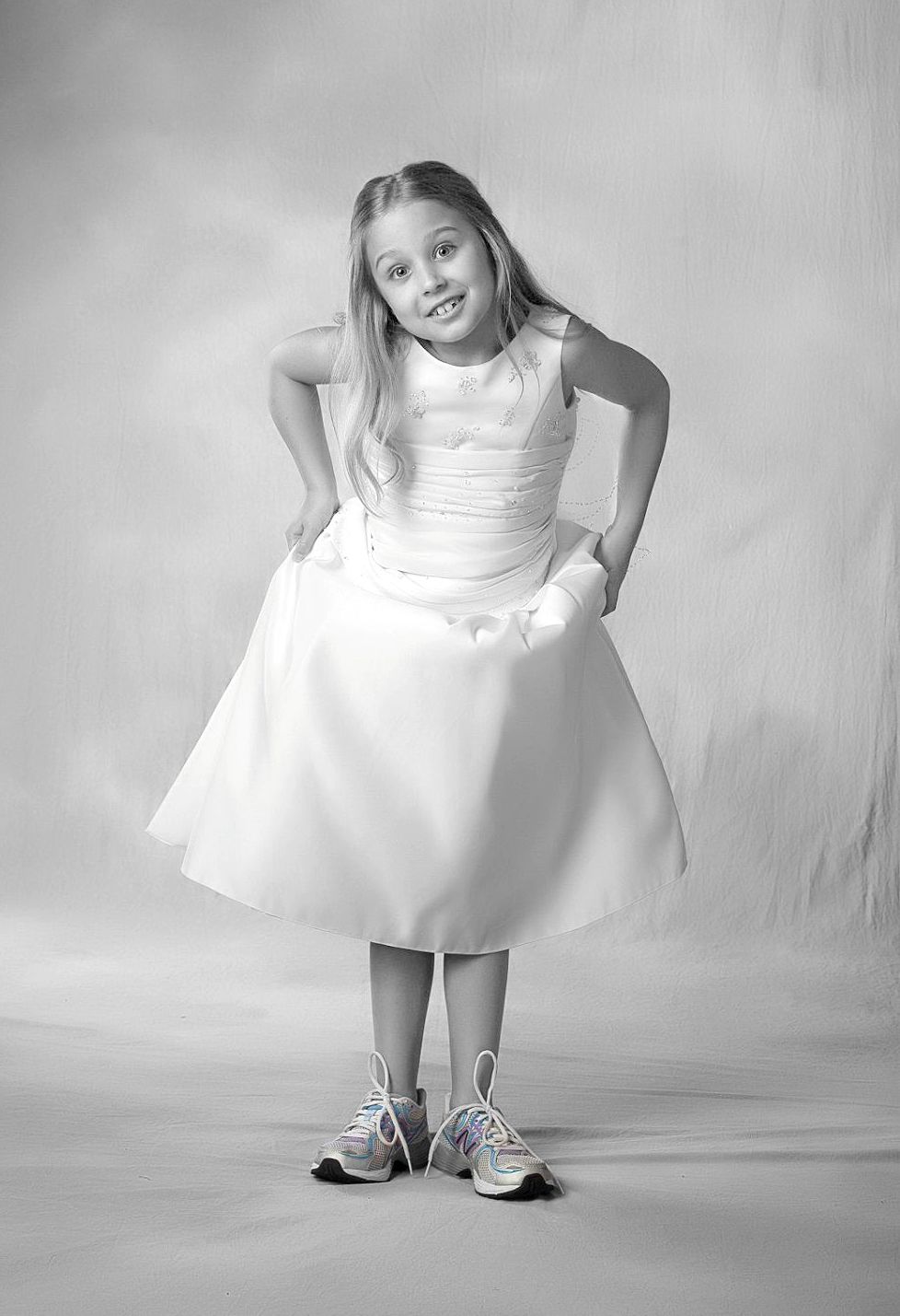 Young girl in white flower girl dress and sneakers posing with hands on hips in black and white portrait.