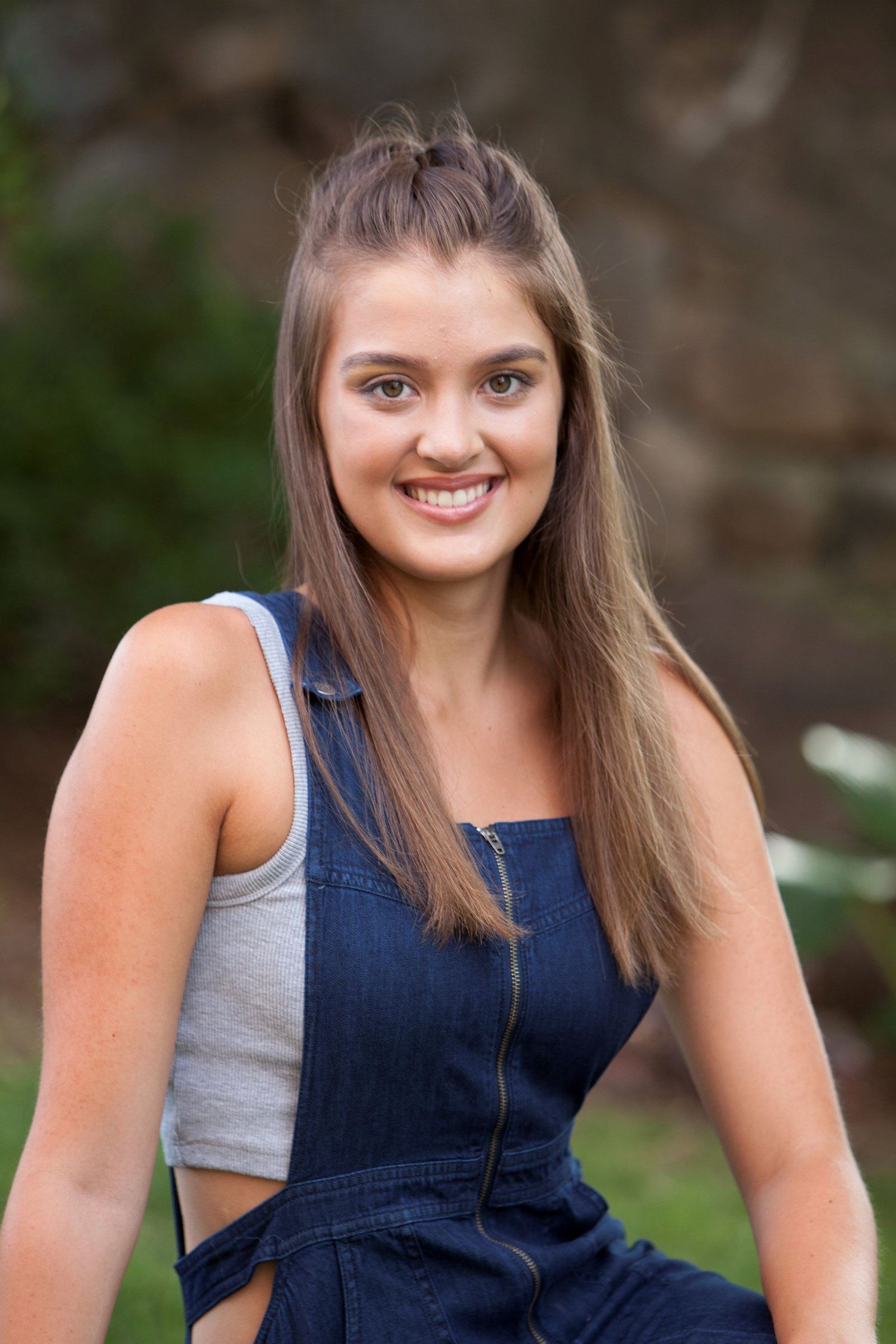 Smiling teenage girl with long brown hair wearing denim overalls poses outdoors against a natural stone background.
