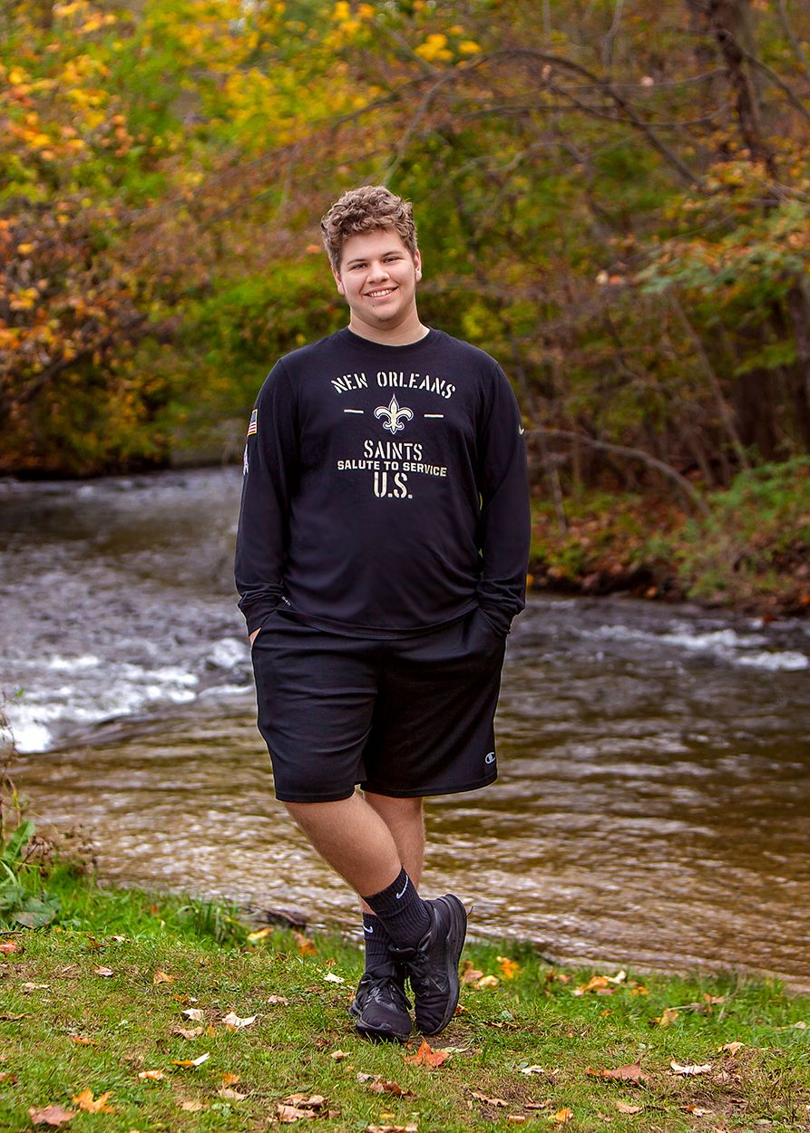 Senior boy in New Orleans Saints shirt standing by a creek in autumn foliage. In Rochester, MI by Jocelyn M. Artistic Photography