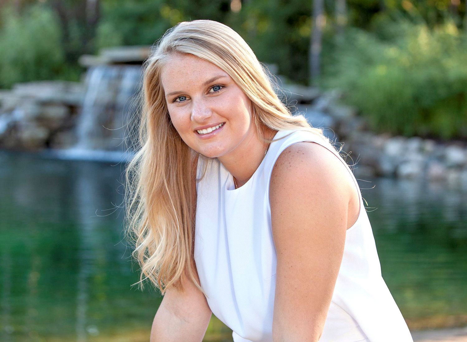 Smiling blonde woman in white sleeveless top posing outdoors near a serene waterfall and pond.