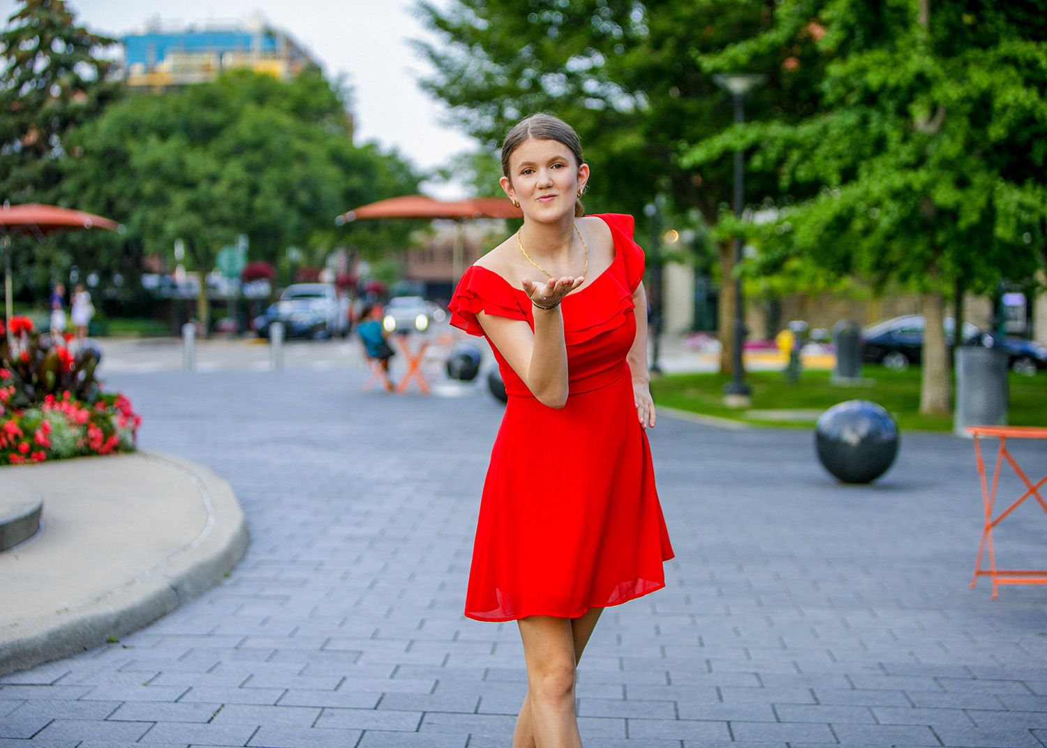 Senior girl blowing a kiss on a sunny urban plaza surrounded by green trees, by Jocelyn M. Photography in Rochester, MI