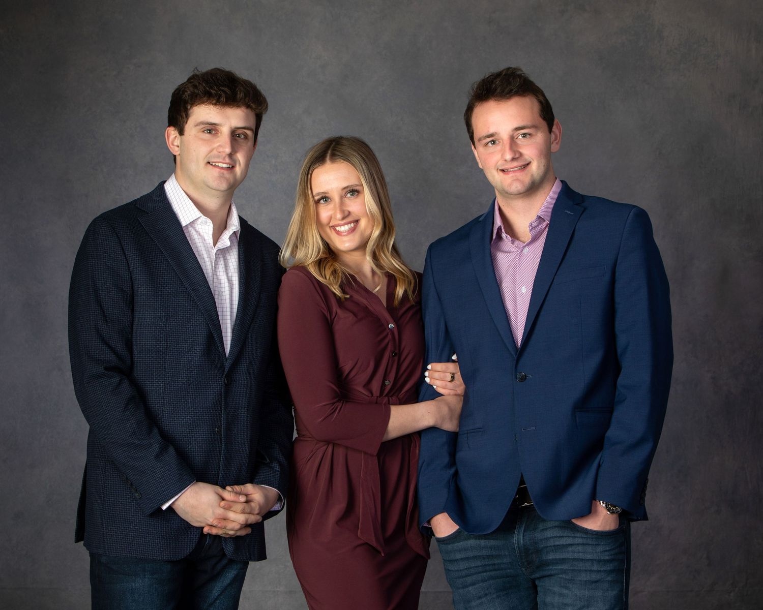Three smiling professionals pose together in business attire against a gray studio backdrop.