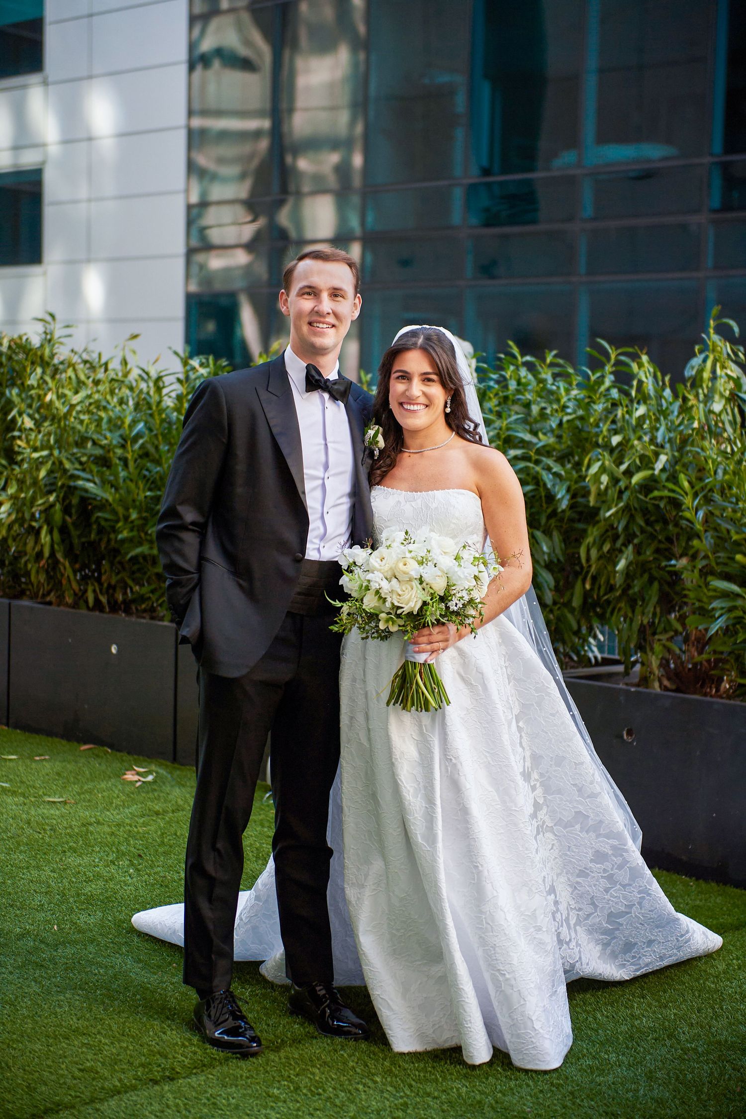 Bride in white strapless gown holding bouquet stands with groom in black tuxedo on green lawn outside modern building.