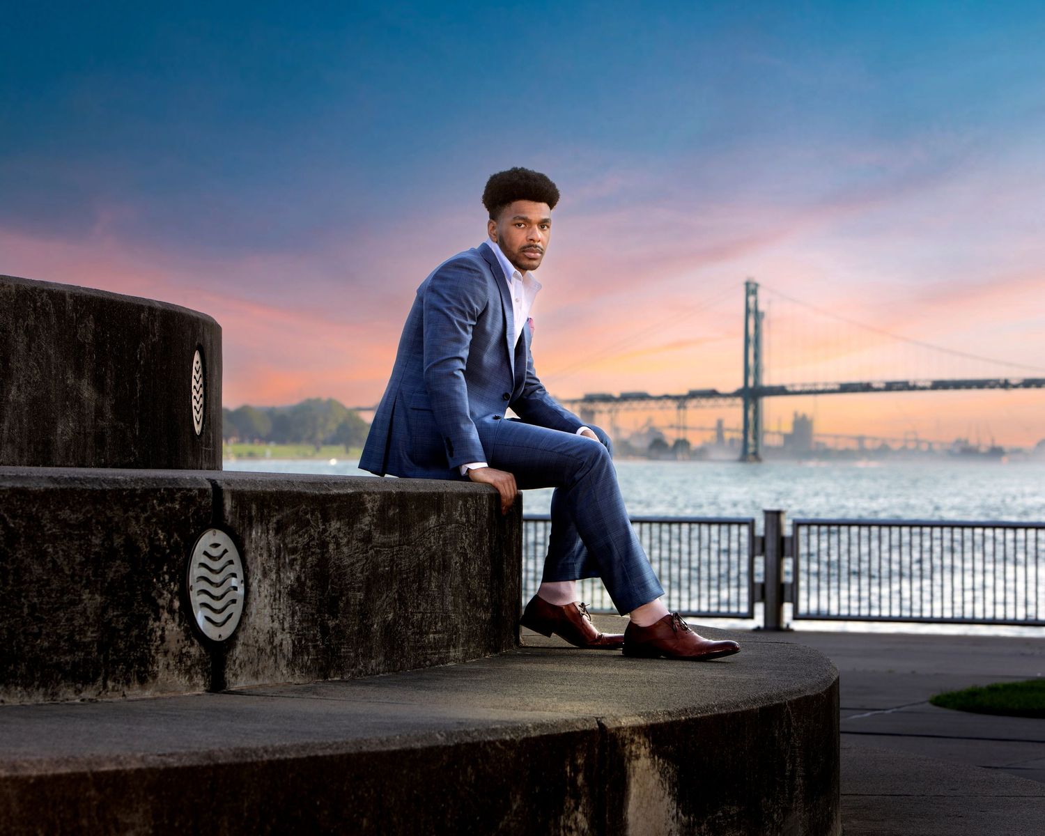 Man in blue suit sitting on stone steps at waterfront with bridge and colorful sunset sky in background.