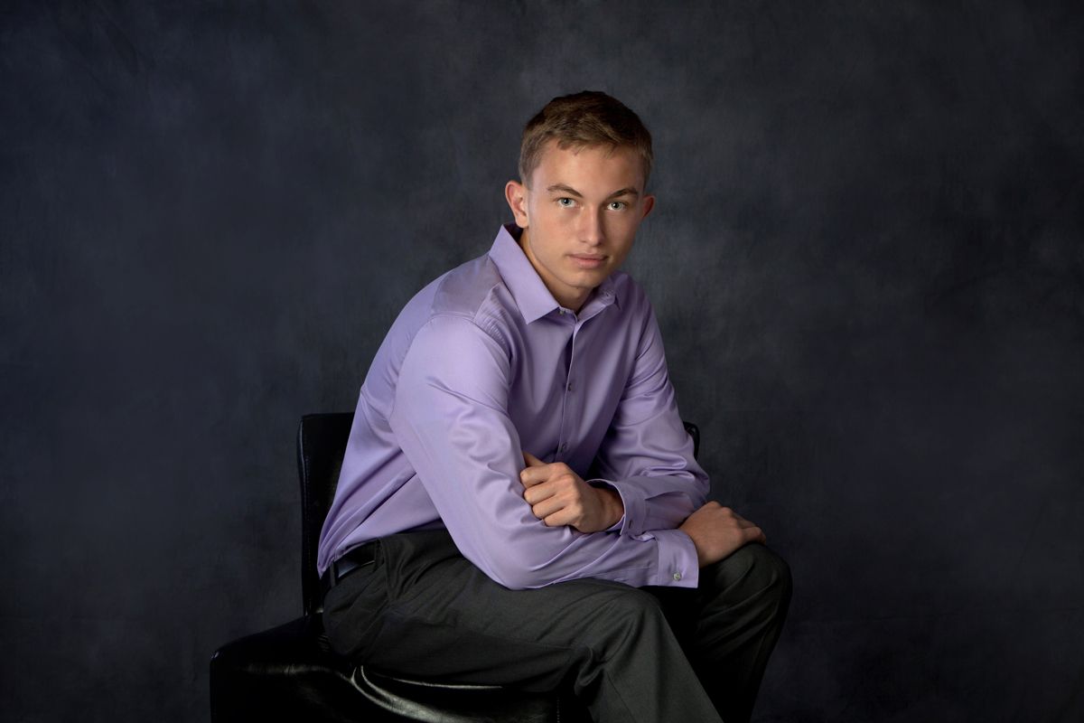 A young man in a purple dress shirt sits confidently on a chair against a dark studio background.