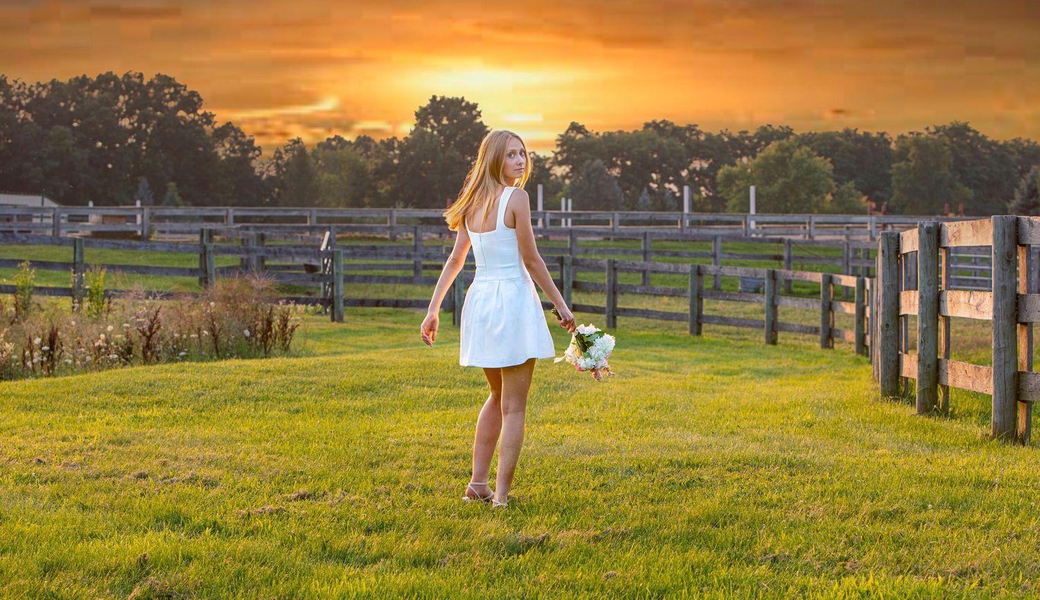 A young woman in a white dress walks through a green farm field at golden sunset, holding a bouquet of flowers.