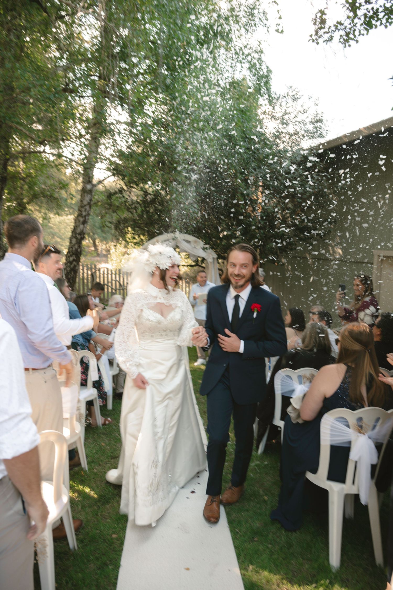 Newlywed couple walks down outdoor aisle as guests toss white confetti during sunny wedding ceremony.
