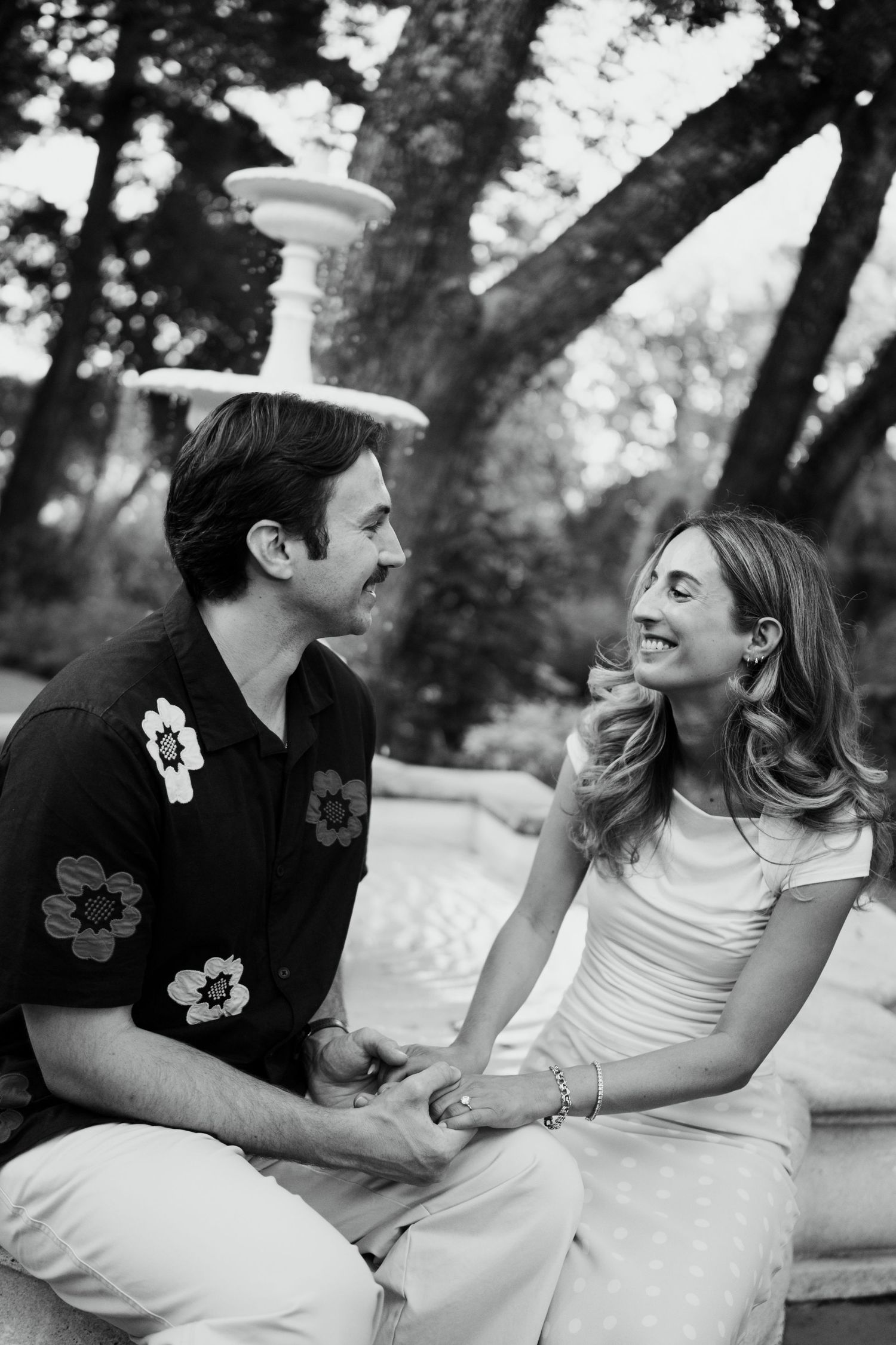 Black and white photo of couple sitting outdoors, smiling at each other near a stone fountain.