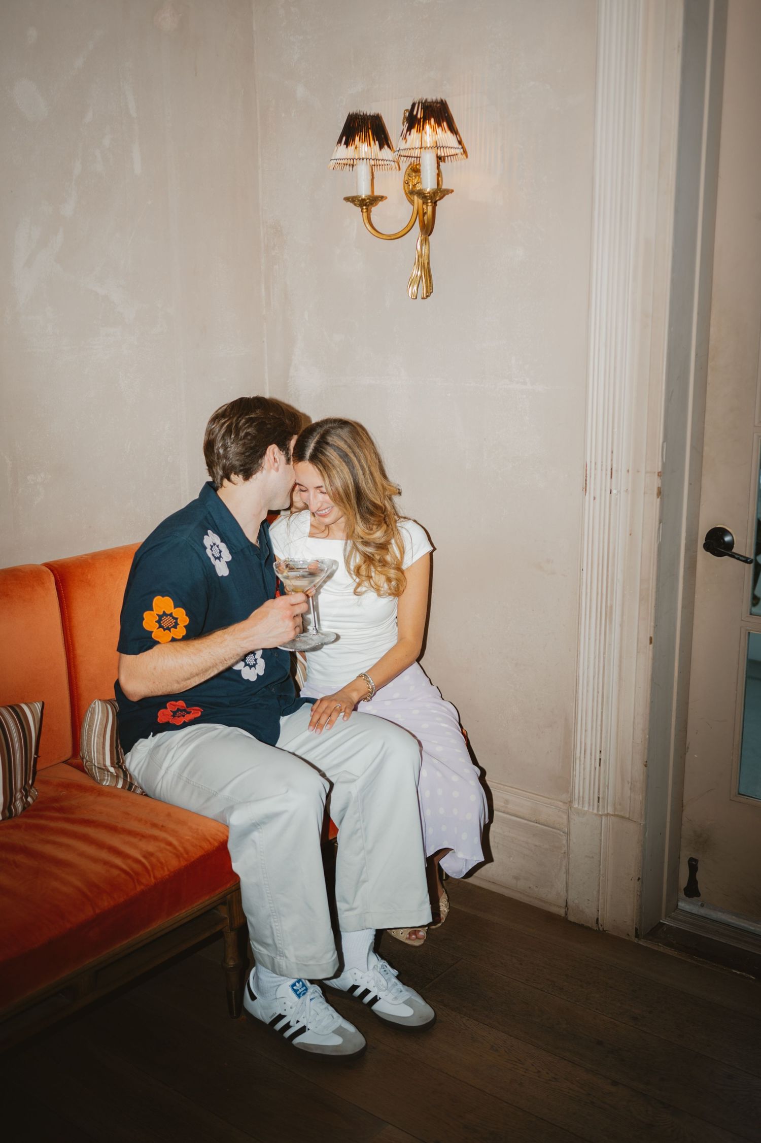 Couple sharing an intimate moment on an orange velvet sofa, holding cocktails beneath a gold wall sconce.