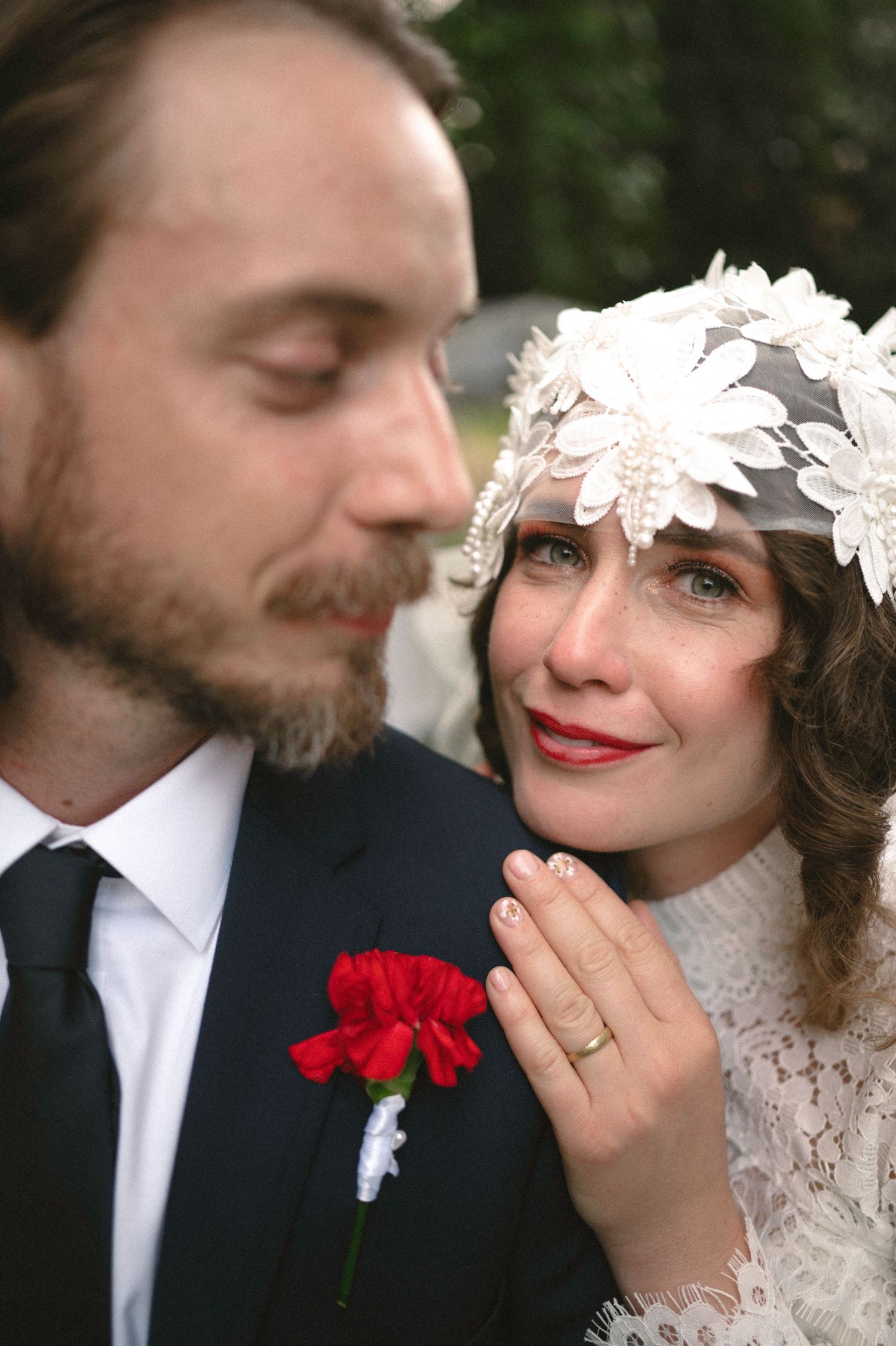 Emotional bride in lace veil and red lips rests head on groom's shoulder at wedding ceremony.