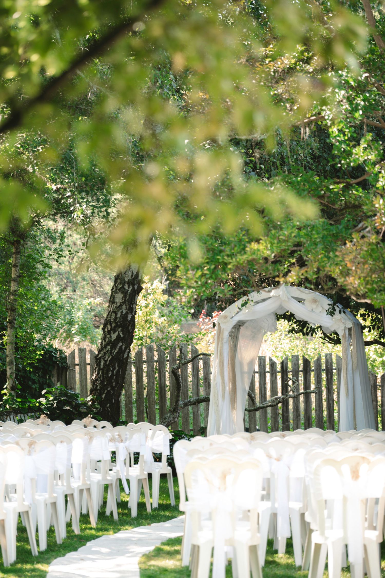 Outdoor wedding ceremony setup with white chairs, floral arch, and lush green trees in a garden setting.