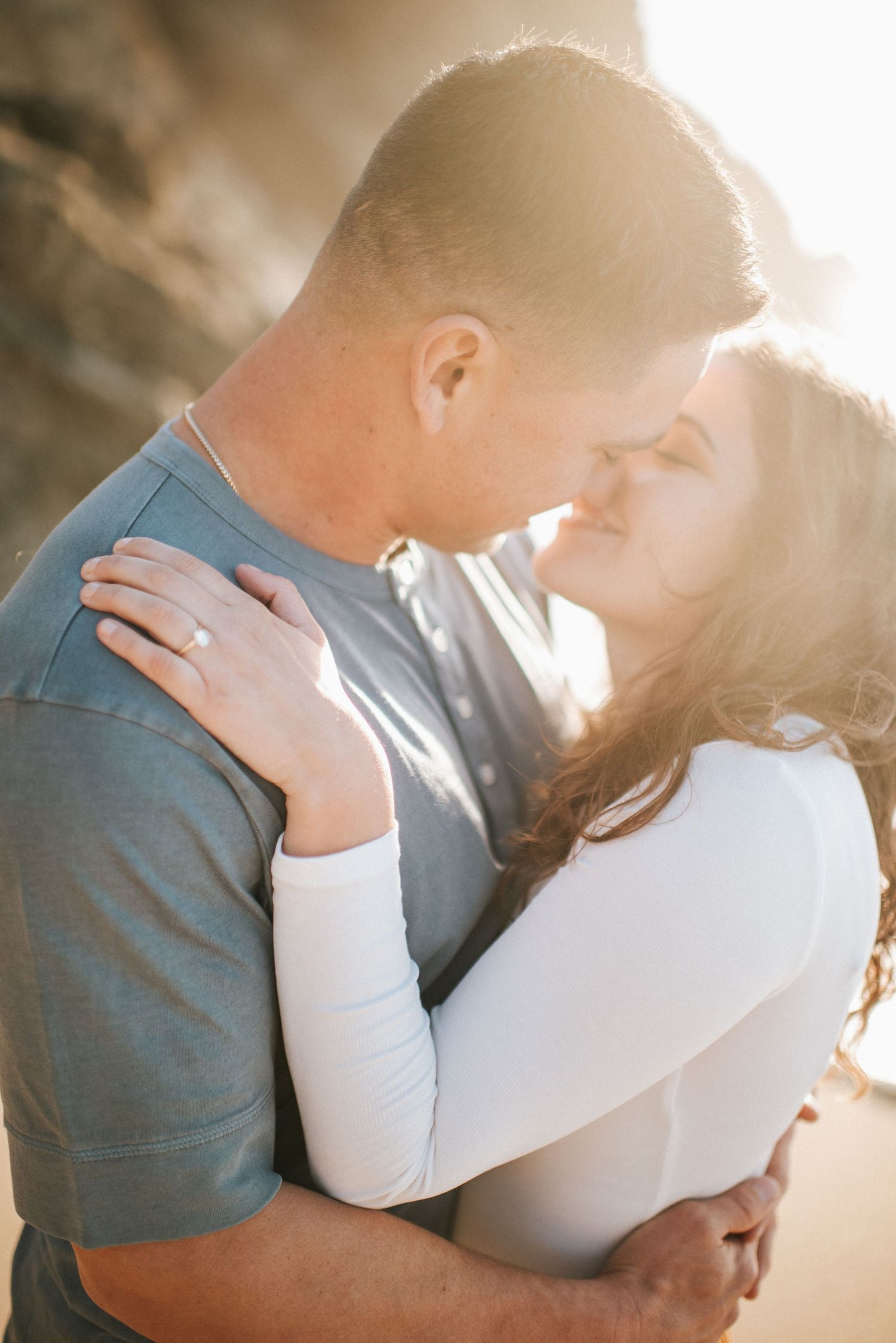 Couple sharing a kiss backlit by golden sunlight, woman wearing an engagement ring.