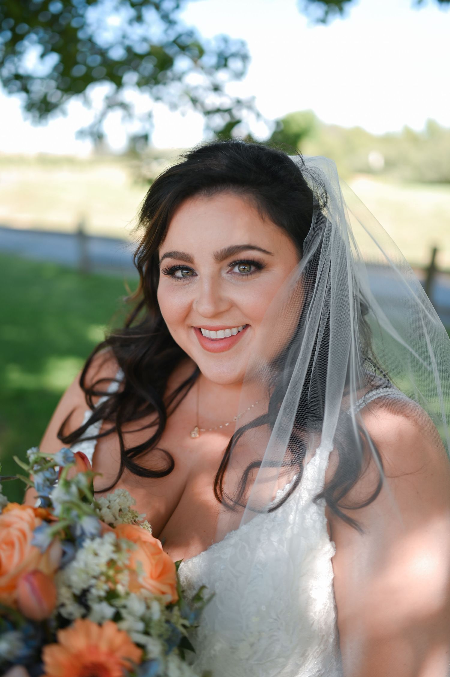Smiling bride in white gown and veil holds peach floral bouquet outdoors on her wedding day.