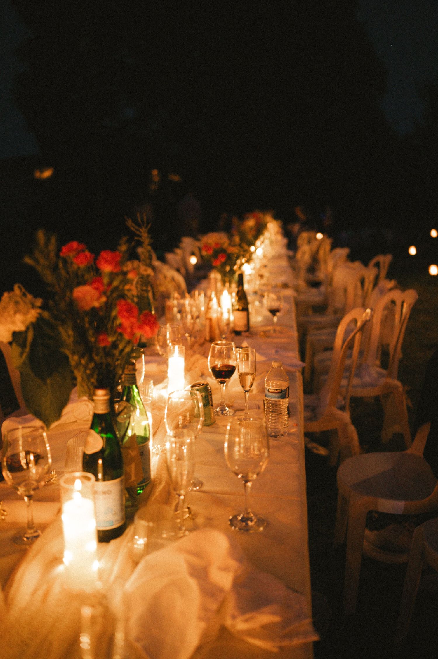 Elegant candlelit outdoor dinner table set with wine glasses, floral centerpieces, and bottles for a romantic evening event.
