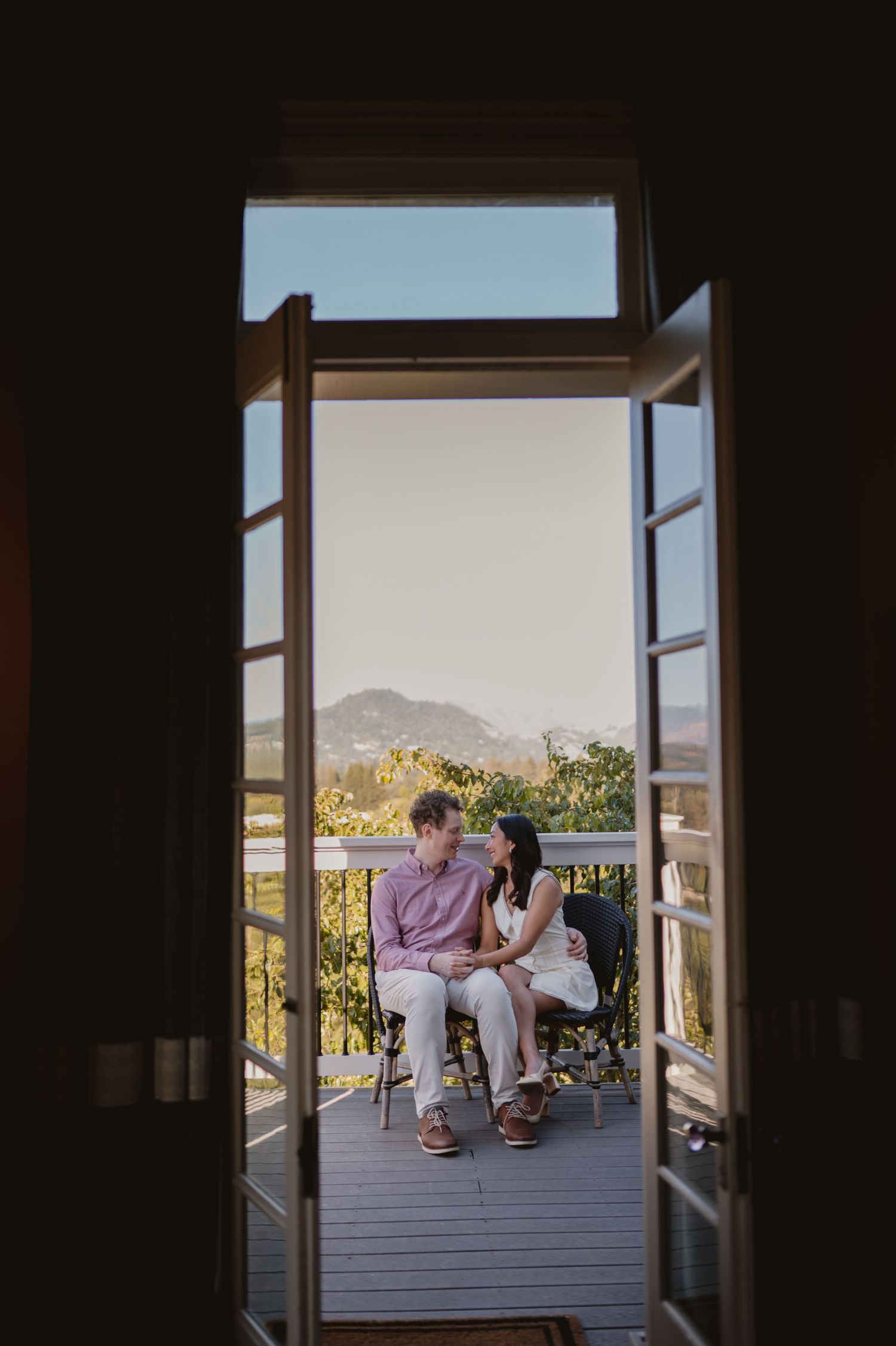 Couple sitting together on a balcony with scenic mountain and vineyard views, framed by open French doors.