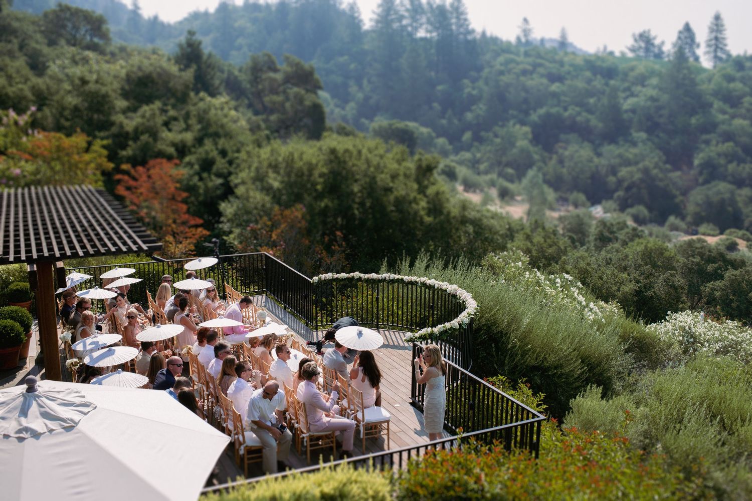 Aerial view of wedding guests dining on a terrace with white umbrellas overlooking lush green forested hills in Sonoma County wine country.