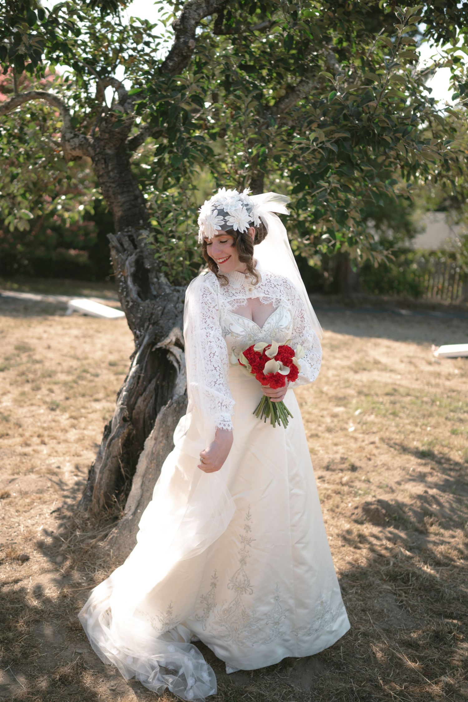Bride in white lace gown and feathered headpiece holds red and white bouquet beside a tree outdoors.