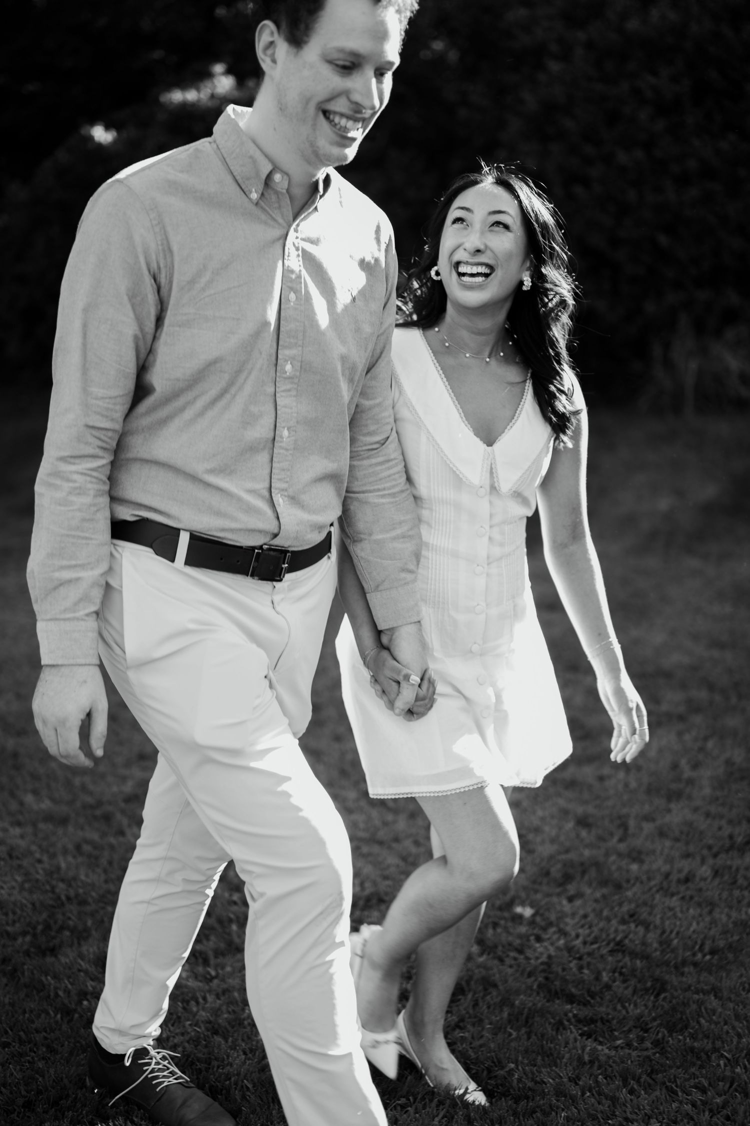 Black and white photo of a happy couple holding hands and walking on grass, woman in white dress laughing joyfully.