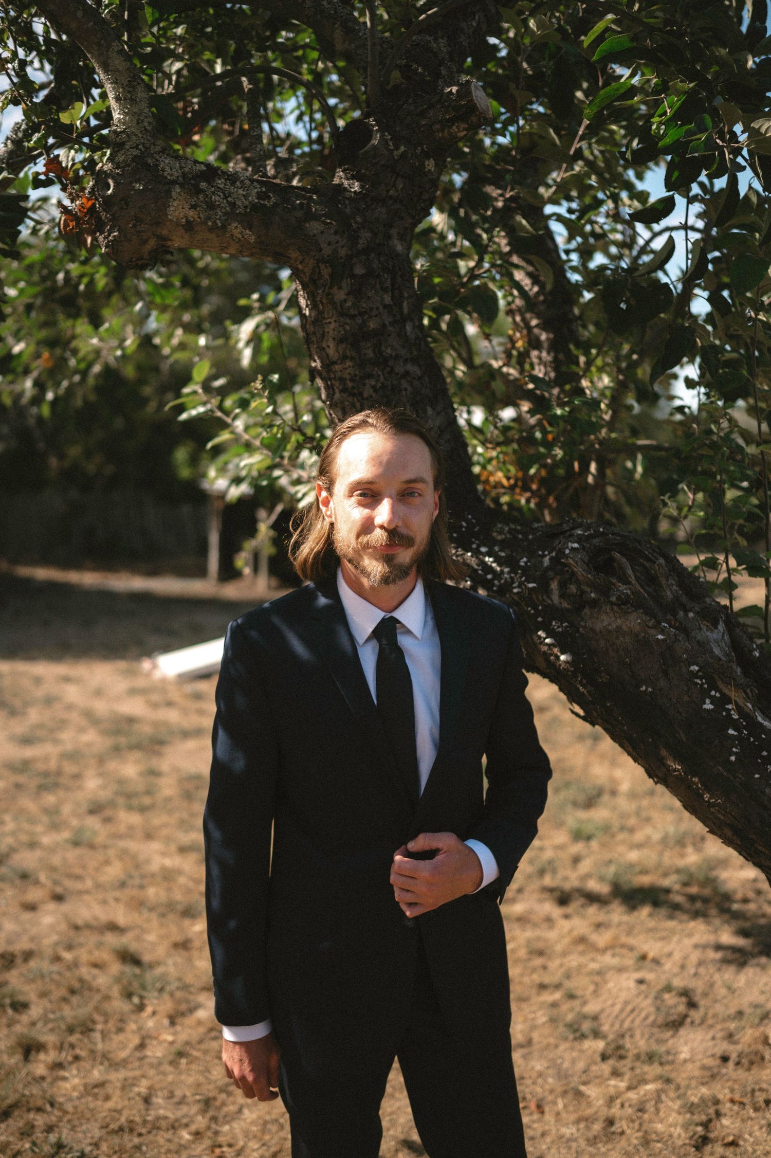 A man with long hair and beard wearing a black suit and tie stands outdoors near a tree.
