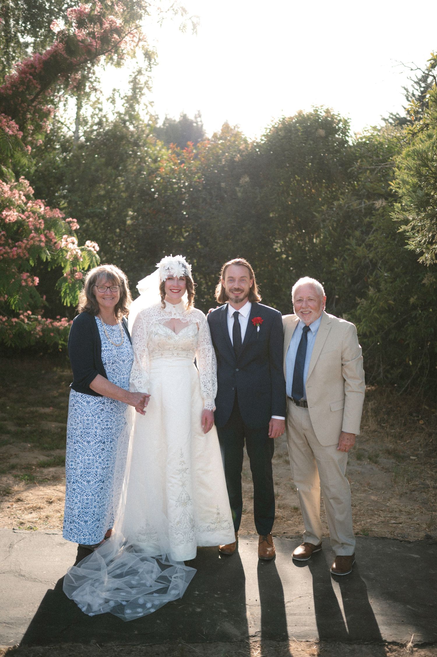 Wedding party of four posing outdoors, bride in lace gown with veil, groom in dark suit, flanked by family members.
