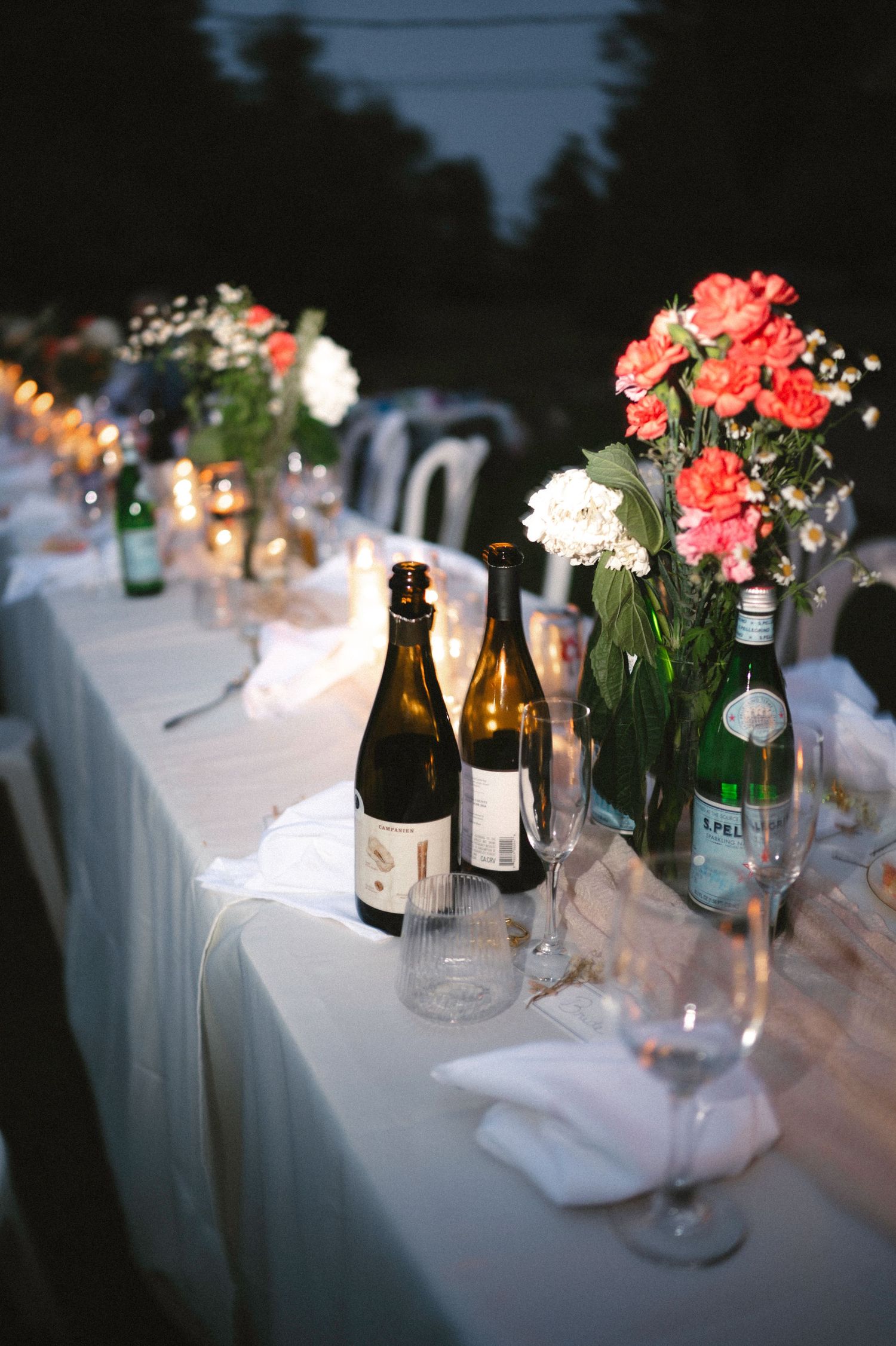 Elegant outdoor wedding reception table with coral flowers, wine bottles, candles, and glassware set on white linen at night.