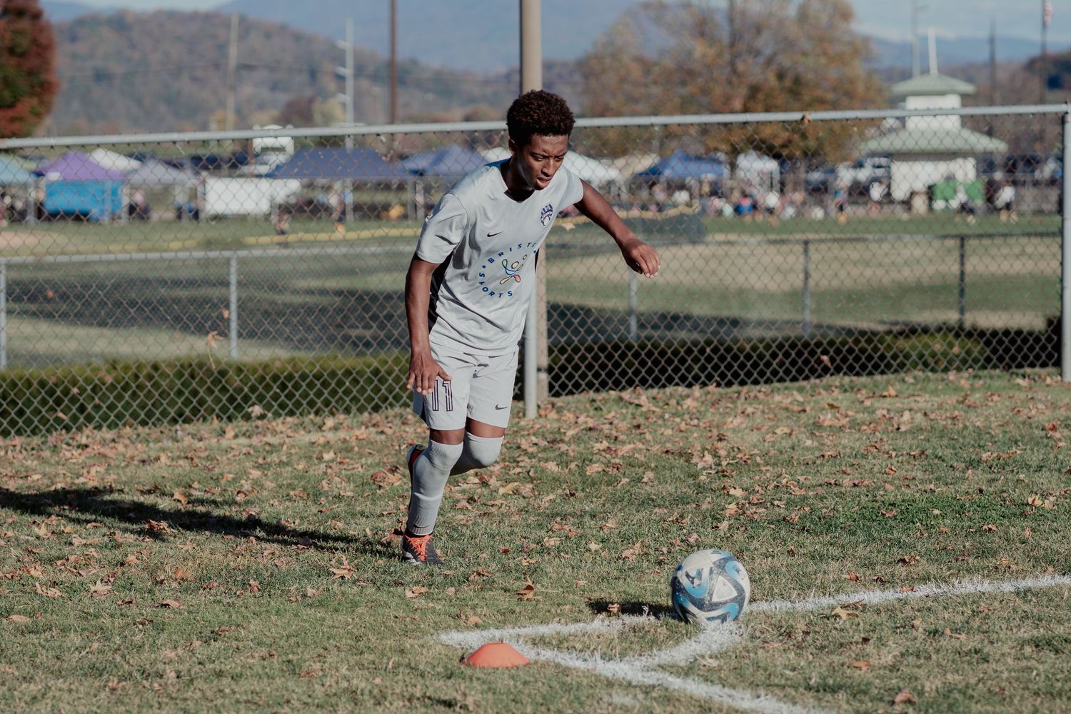 A soccer player in white uniform prepares to kick a ball on a dry grass field with mountains in the background.