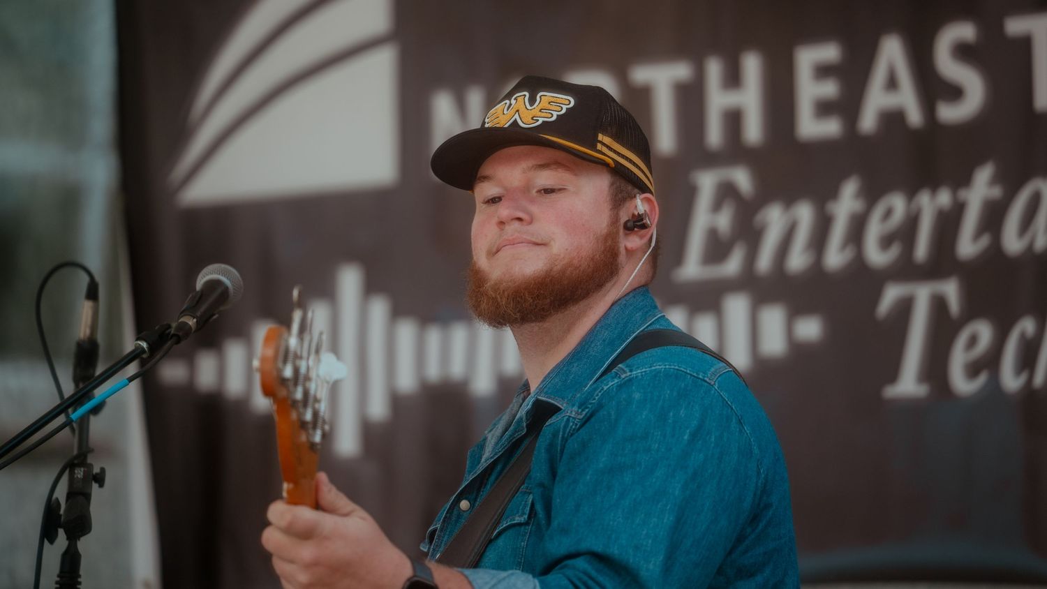 A musician in a blue button-down shirt performs on stage with a bass guitar at a North East Entertainment event.