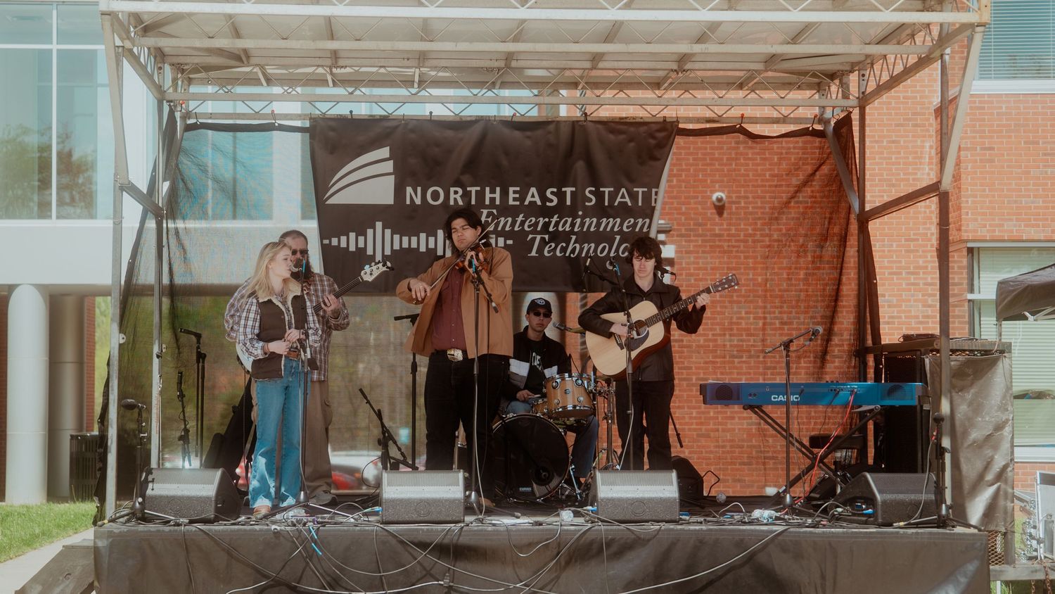 Musicians perform on an outdoor stage setup under a canopy with brick wall background during a live concert event.