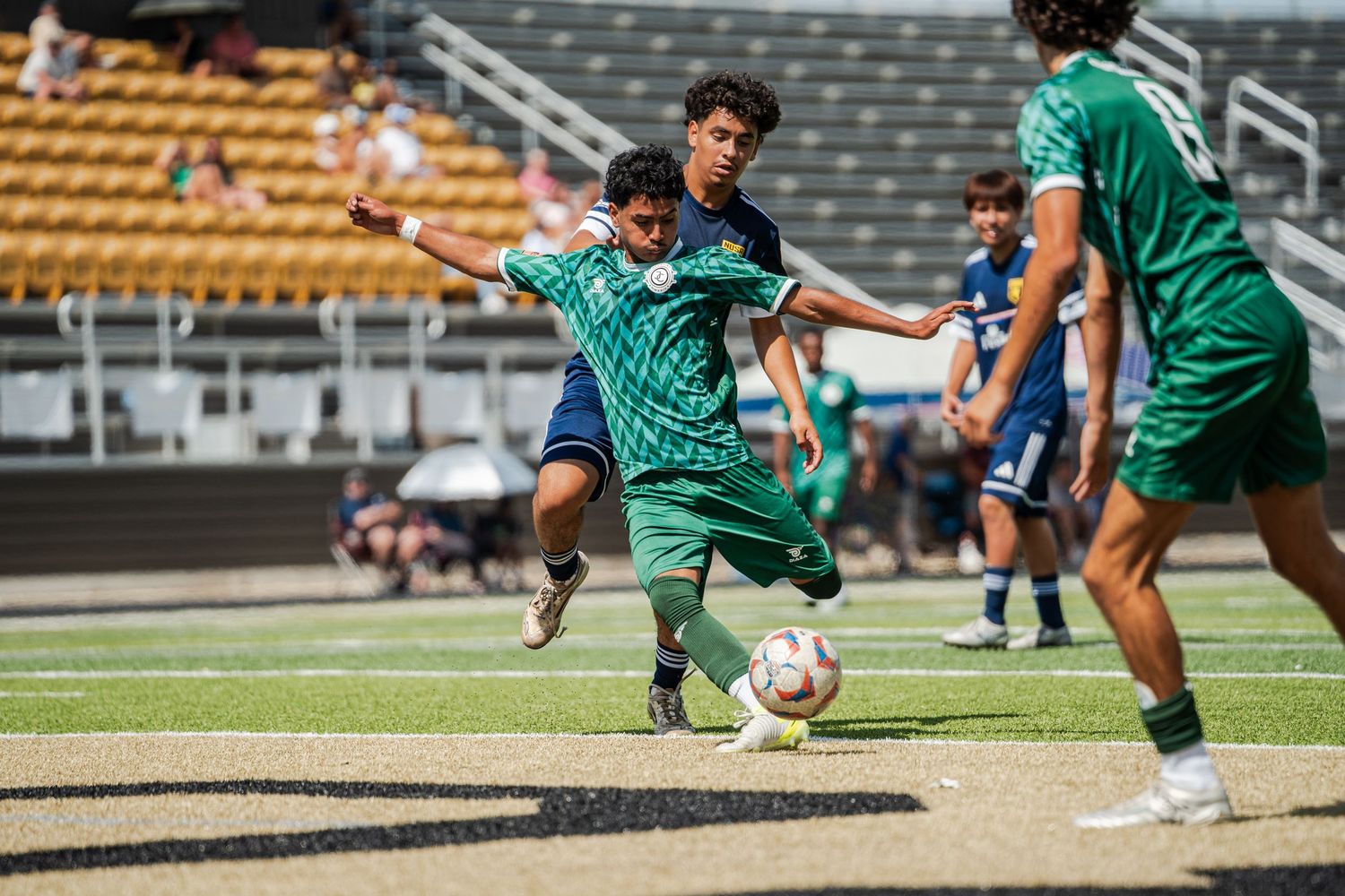 Two soccer players in green uniforms compete for the ball during a sunny outdoor match on a grass field.
