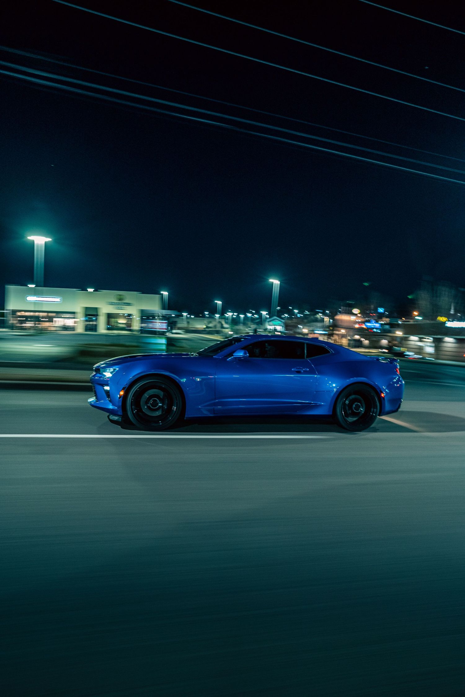 A bright blue Chevrolet Camaro sports car drives at night through a dimly lit parking lot with street lights overhead.
