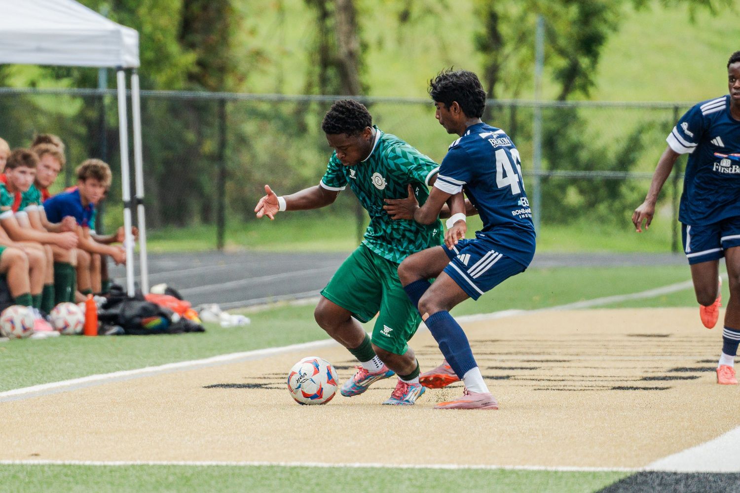 Two youth soccer players compete for the ball during a match, one in green and one in navy blue uniforms.