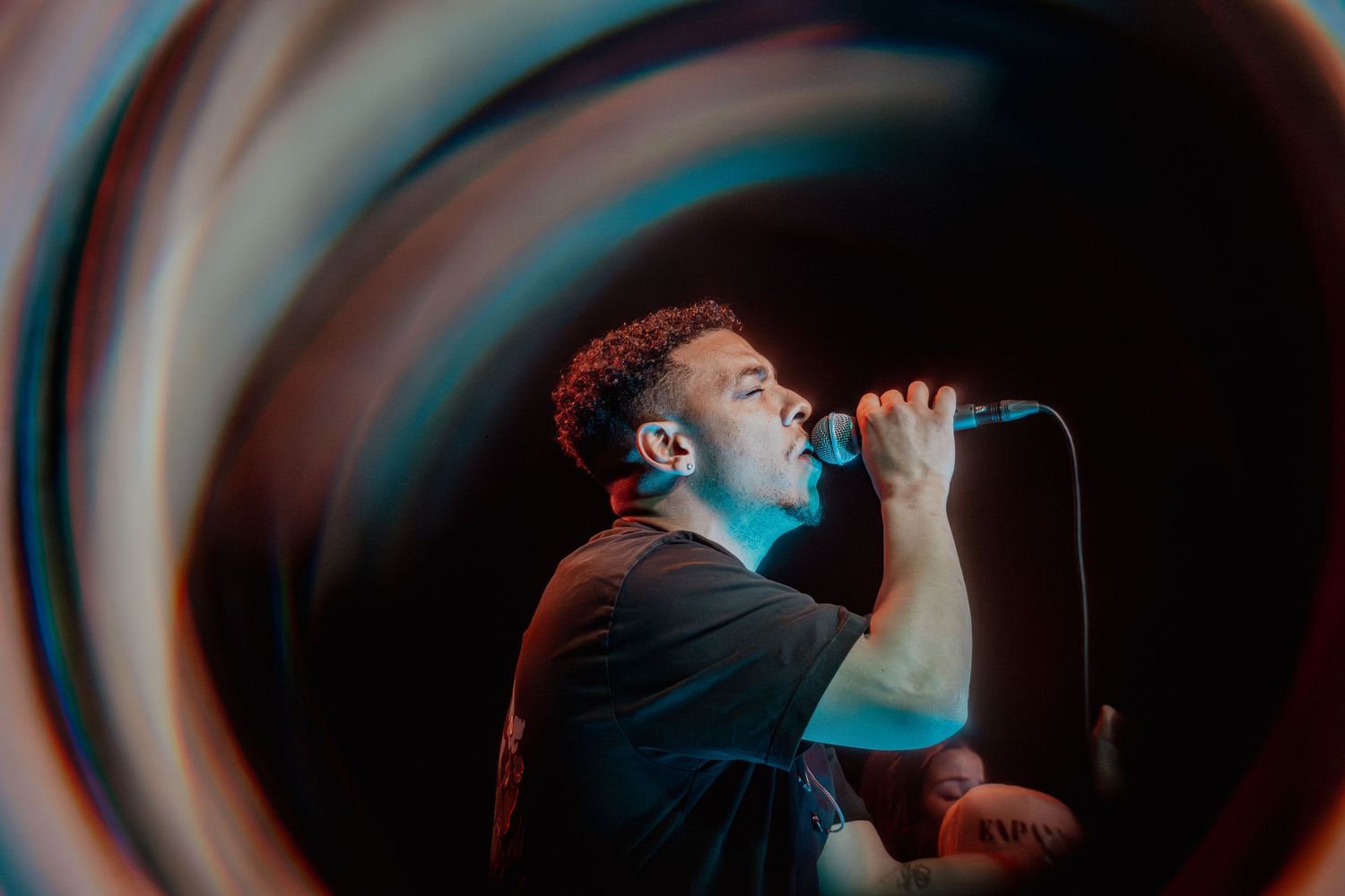 A male vocalist singing passionately into a microphone on stage, lit by blue and warm lights with a dramatic vignette effect.