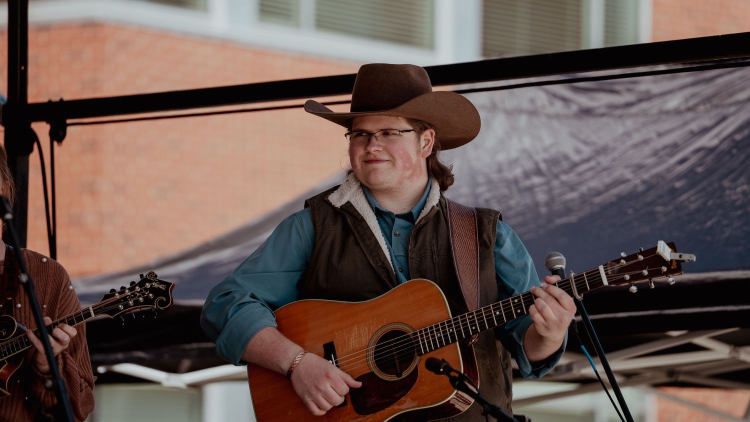 A country musician performs on stage with an acoustic guitar while wearing a brown cowboy hat and blue denim jacket.