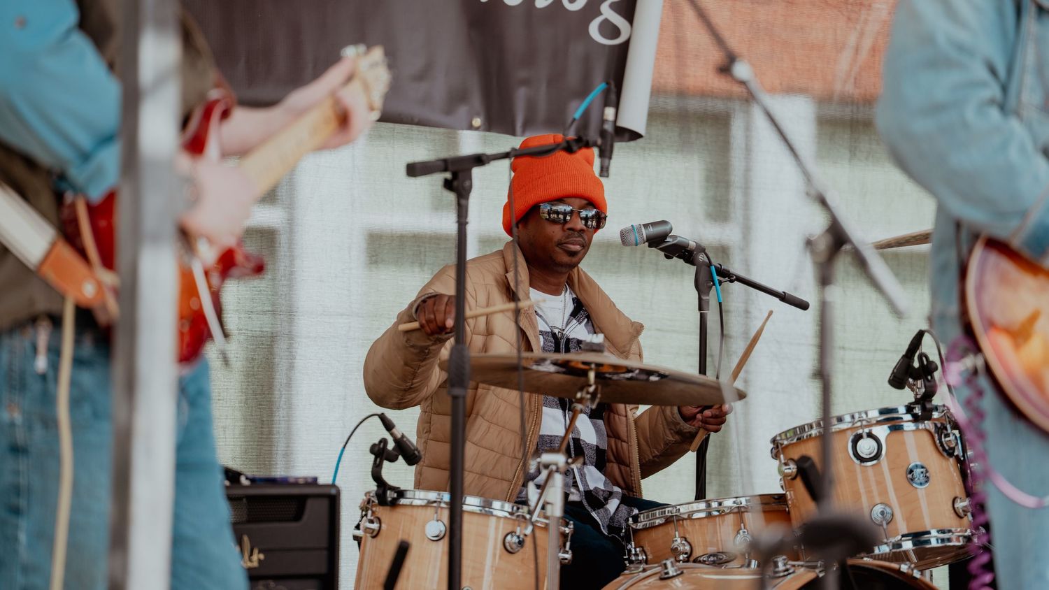 A drummer wearing a orange beanie and sunglasses performs energetically behind a drum kit on stage at an outdoor event.