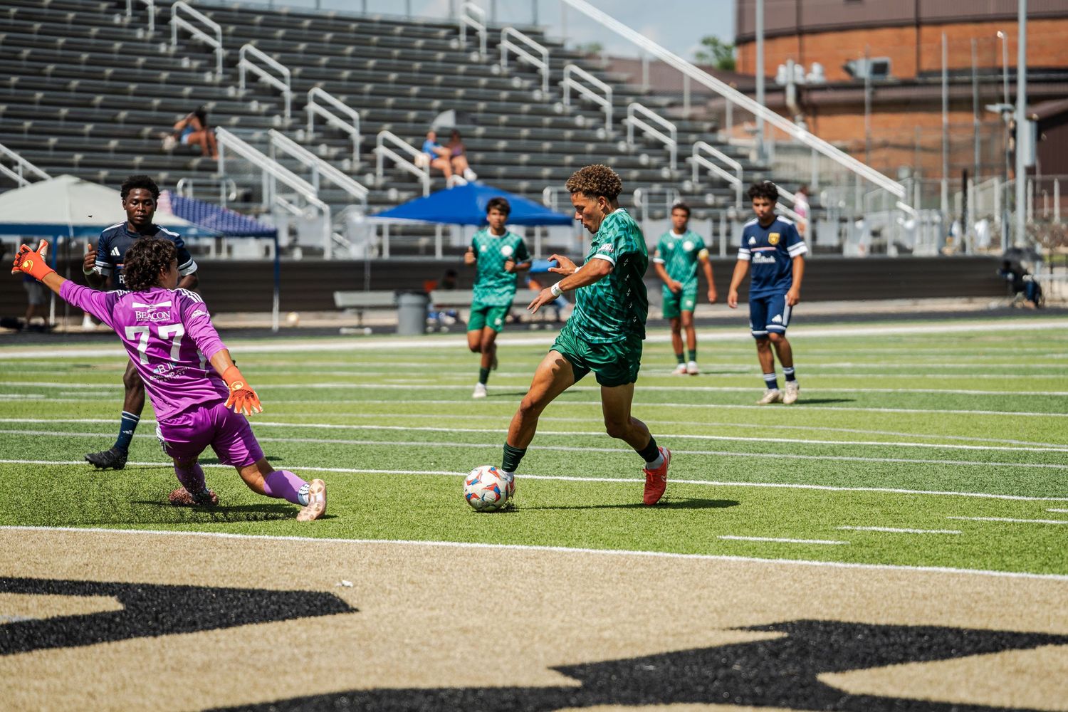 Youth soccer player in green dribbles past a diving goalkeeper in purple during a sunny outdoor match.