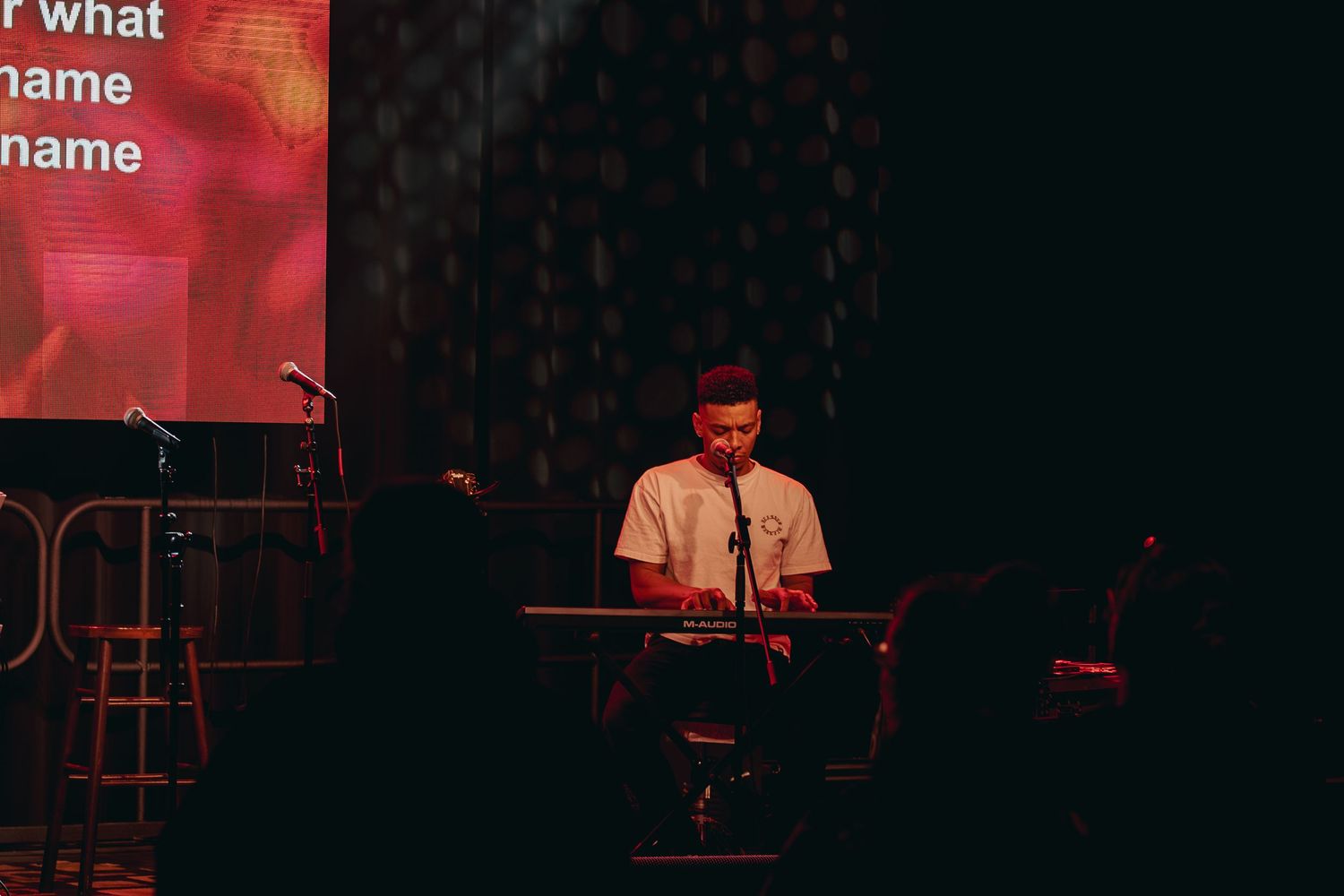 A performer plays electronic music equipment on a dimly lit stage with red lighting in the background.